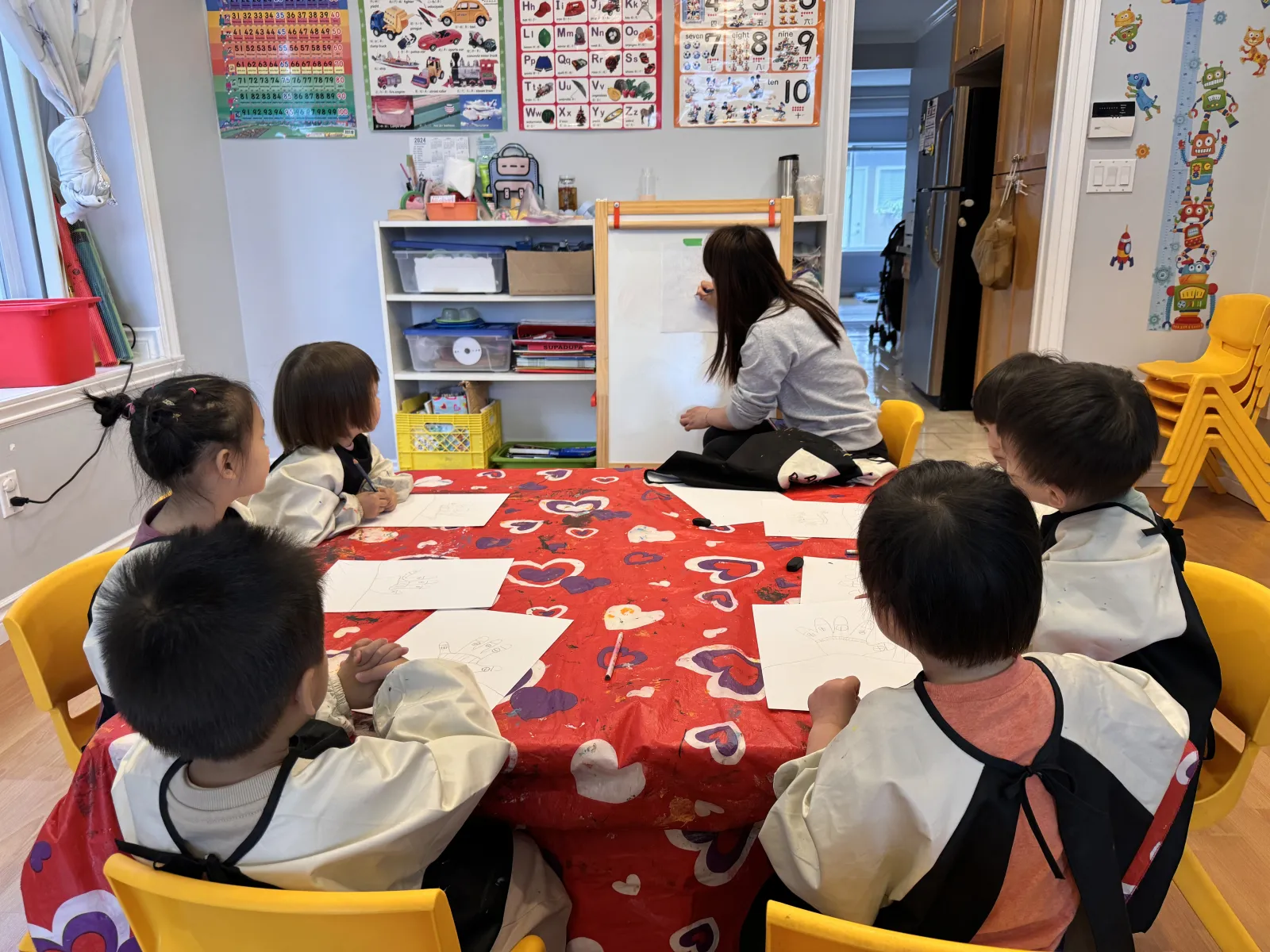 Children drawing at an art table while their teacher sketches on an easel